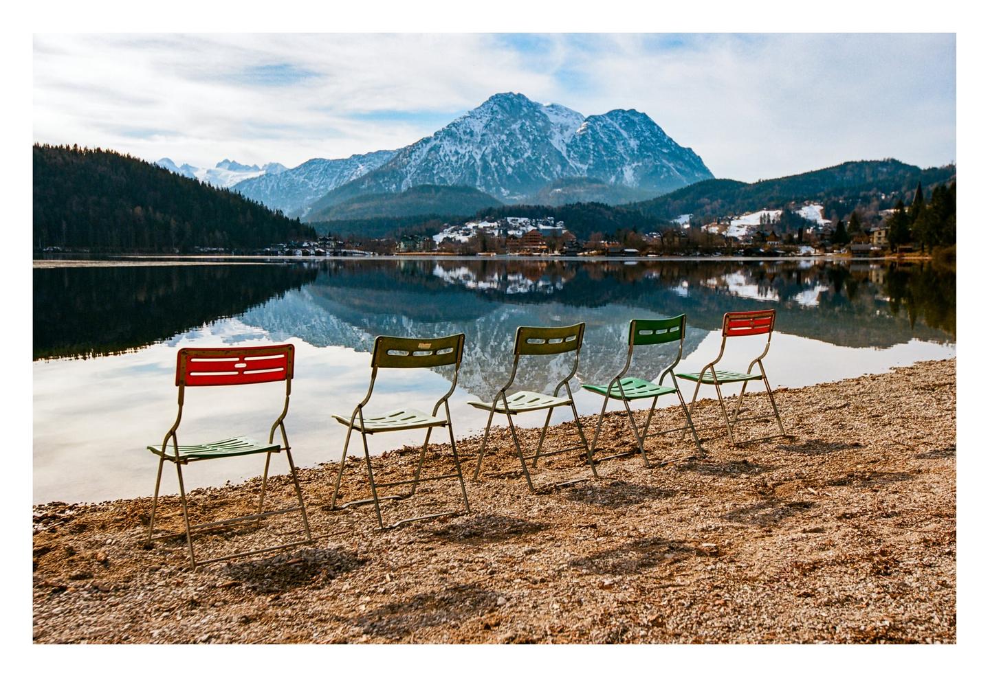 Five chairs sit on a rocky shore overlooking a lake with snow-capped mountains and a village in the background.