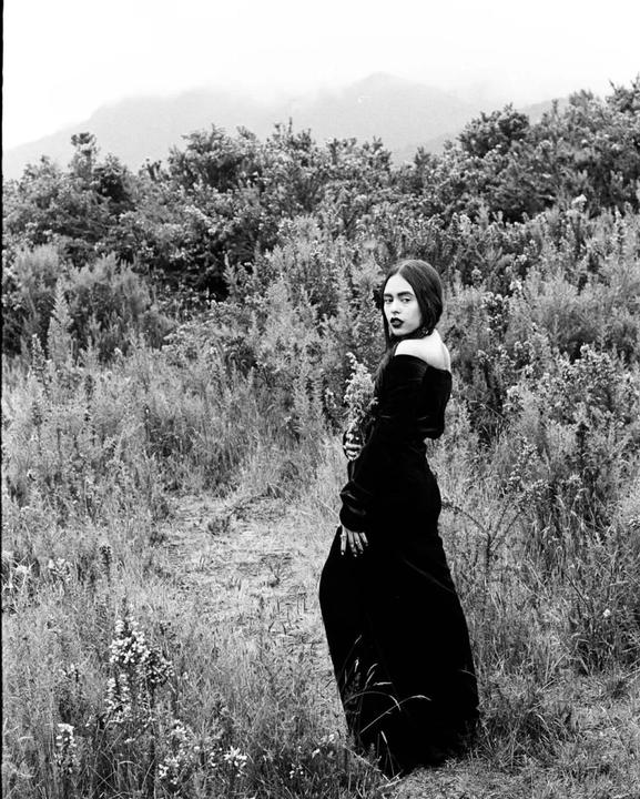 A woman in a dark dress stands in a field of tall grass and shrubs with mountains in the background.