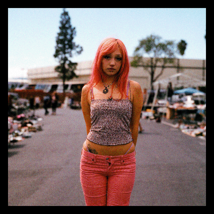 A woman with pink hair and pink pants stands in the middle of an outdoor market.