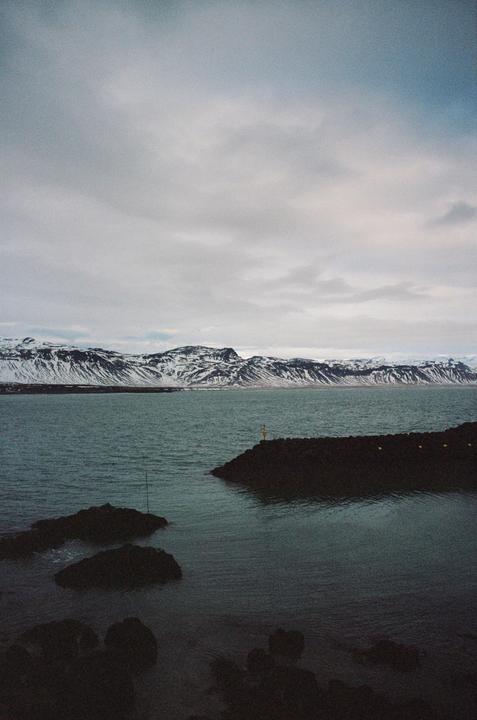 A body of water is in the foreground with snow-capped mountains in the background under a cloudy sky.