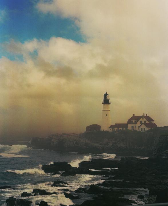 A lighthouse stands on a rocky coast with waves crashing below and a cloudy sky above.