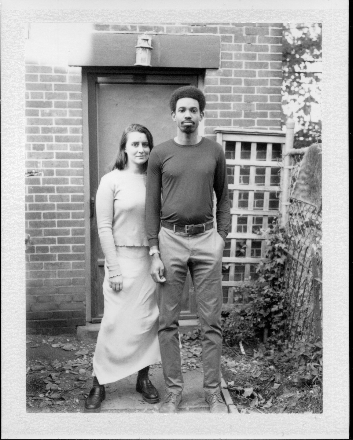 A man and a woman stand in front of a brick wall and a door.