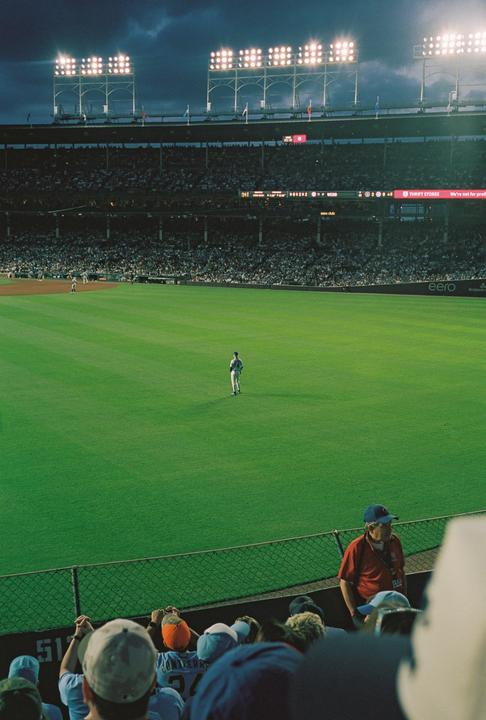 A baseball player stands alone on a green field in a stadium filled with spectators under bright lights at dusk.
