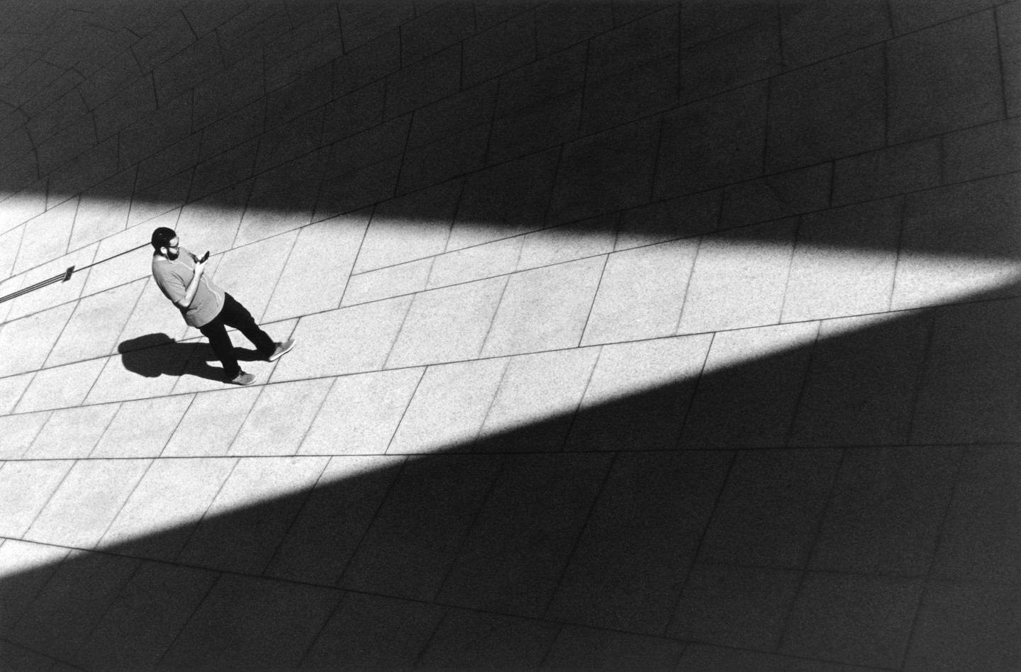 A man stands on a tiled surface, looking at his phone, with a strong diagonal shadow cutting across the frame.