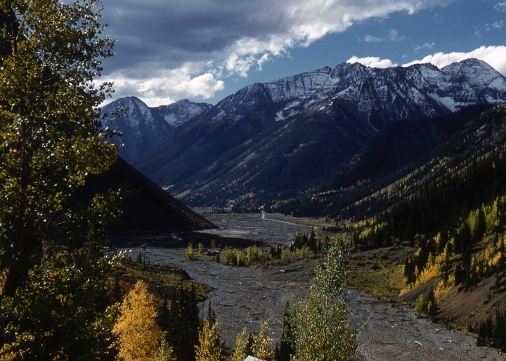 A valley with a riverbed and autumn trees is surrounded by mountains with snow-capped peaks under a cloudy sky.