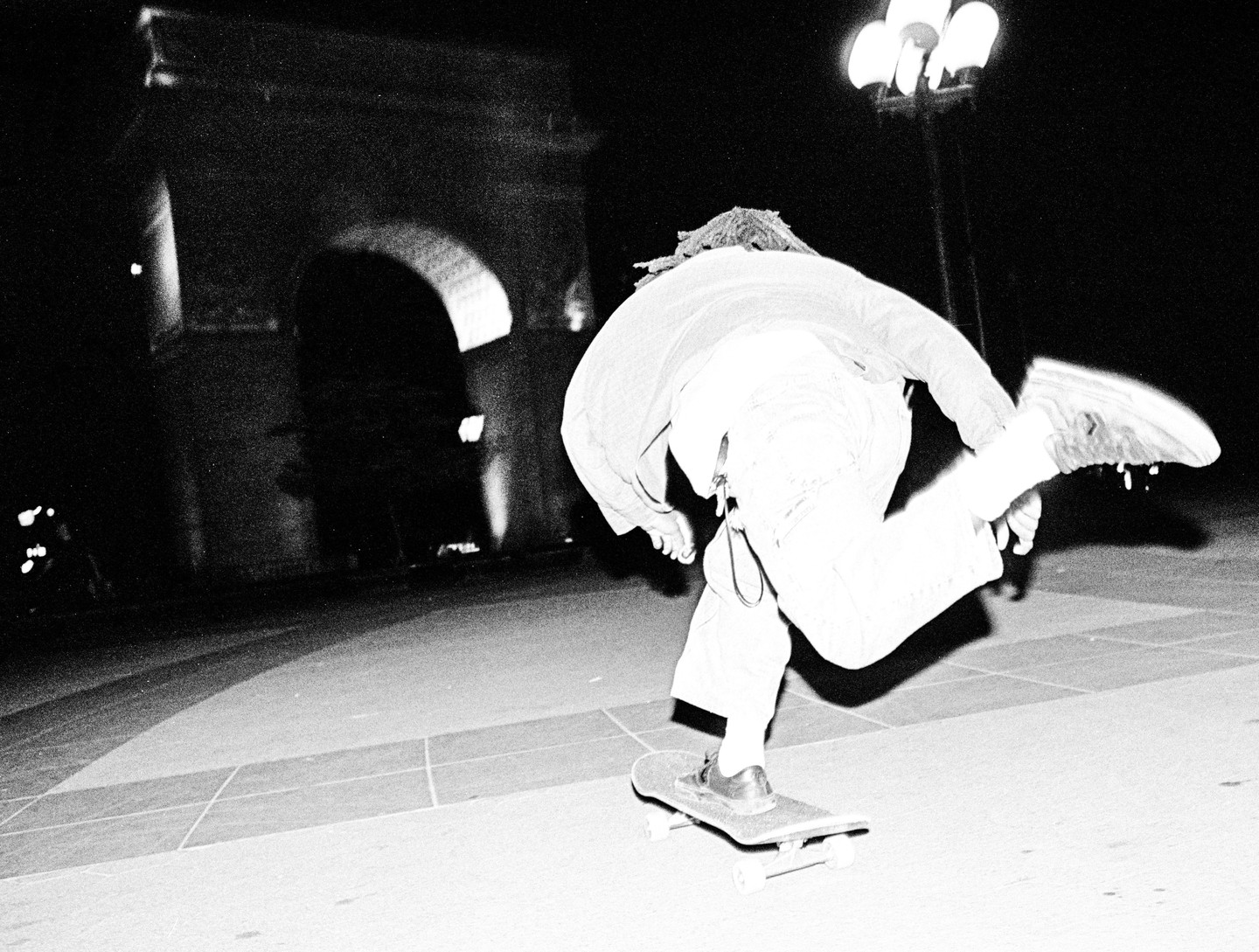 A person on a skateboard is captured in motion at night near an arch and a street light.