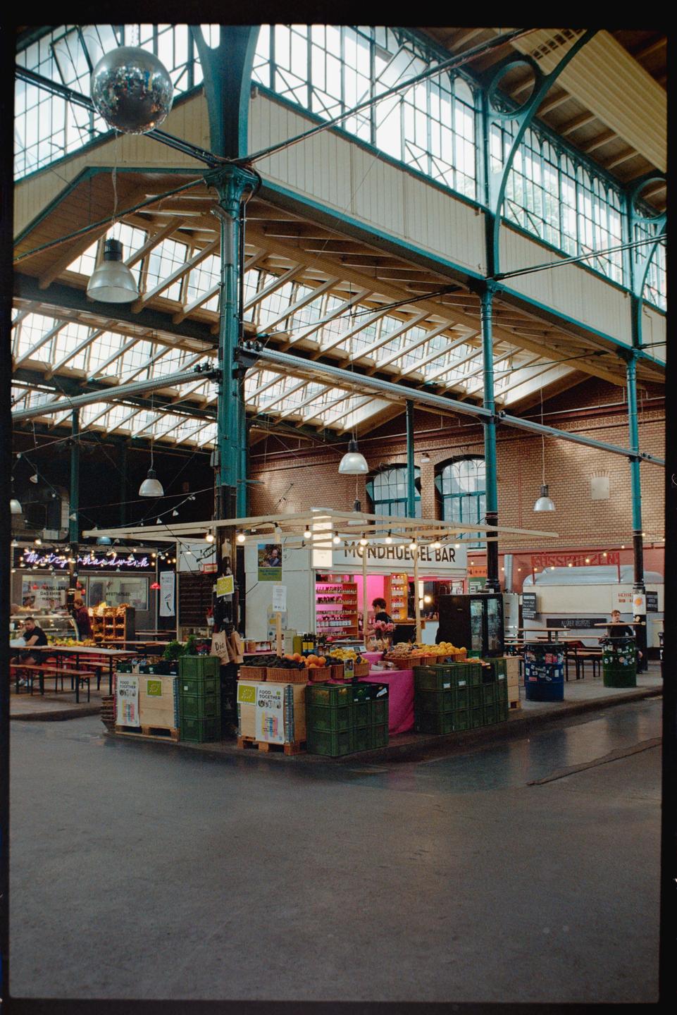 A disco ball hangs from the ceiling of a market hall with various stalls and people.