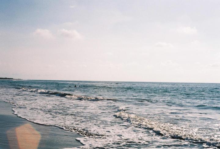 Waves crash on a sandy beach with a few people swimming in the distance under a bright sky.