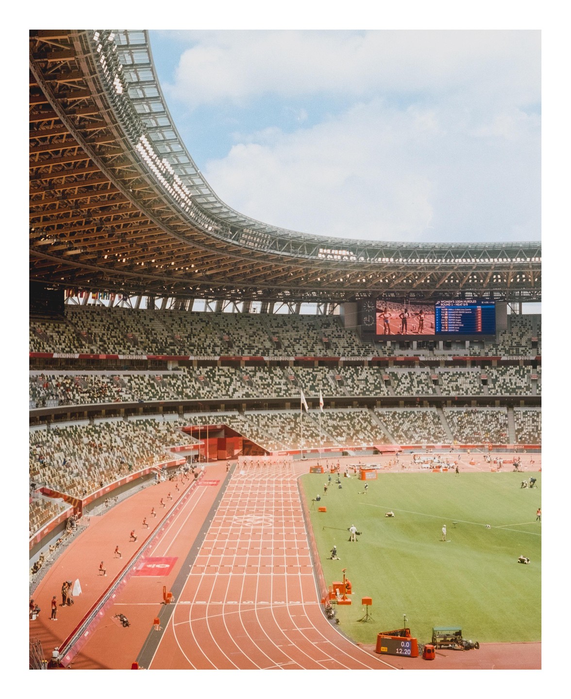 A wide view of the Tokyo Olympic Stadium shows a track and field event in progress with a large screen displaying results.