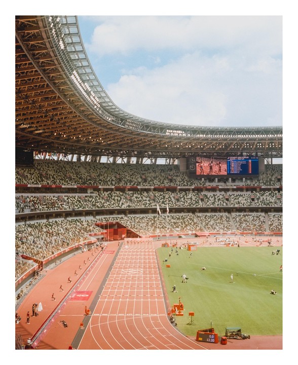 A wide view of the Tokyo Olympic Stadium shows a track and field event in progress with a large screen displaying results.