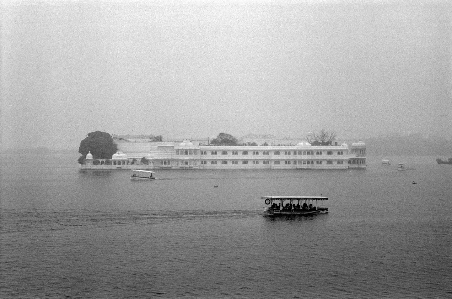 A large white building with many windows sits on an island in the middle of a body of water, with several boats moving across the water.