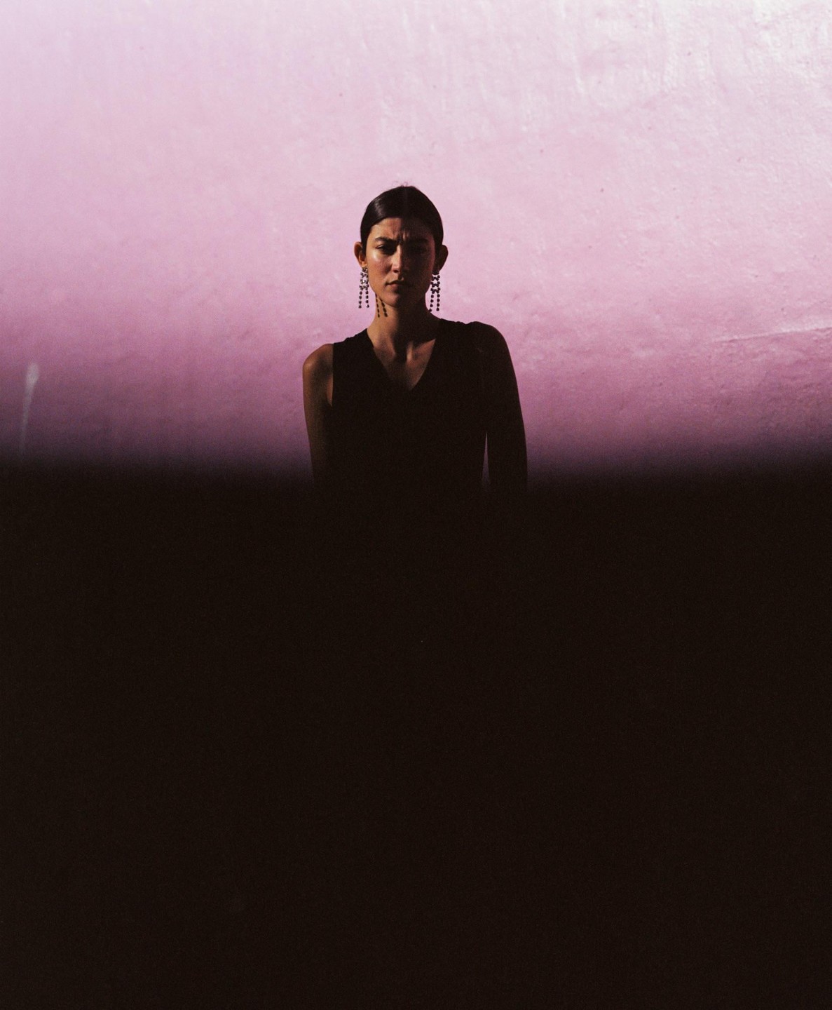 A woman with dark hair and earrings stands in front of a pink wall, with the lower half of the image in shadow.