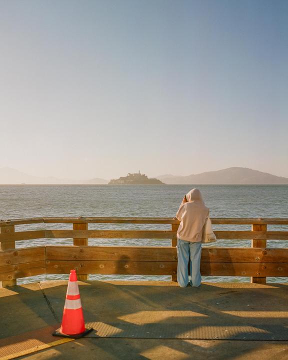 A person in a hoodie stands on a pier looking out at Alcatraz Island in the distance during golden hour.