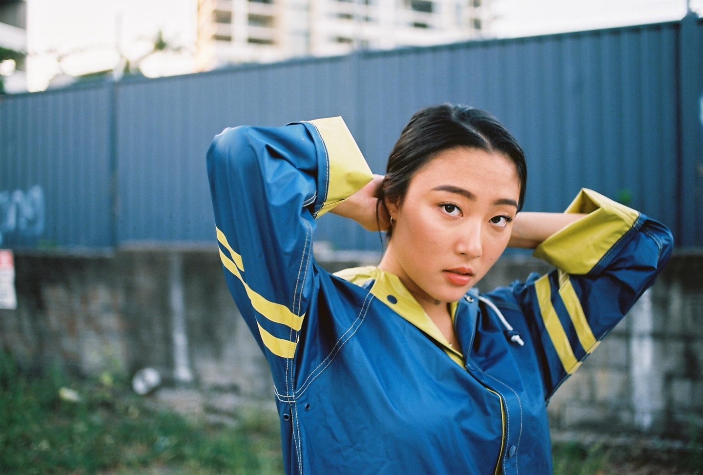 A woman in a blue and yellow jacket poses with her hands behind her head in an urban setting.