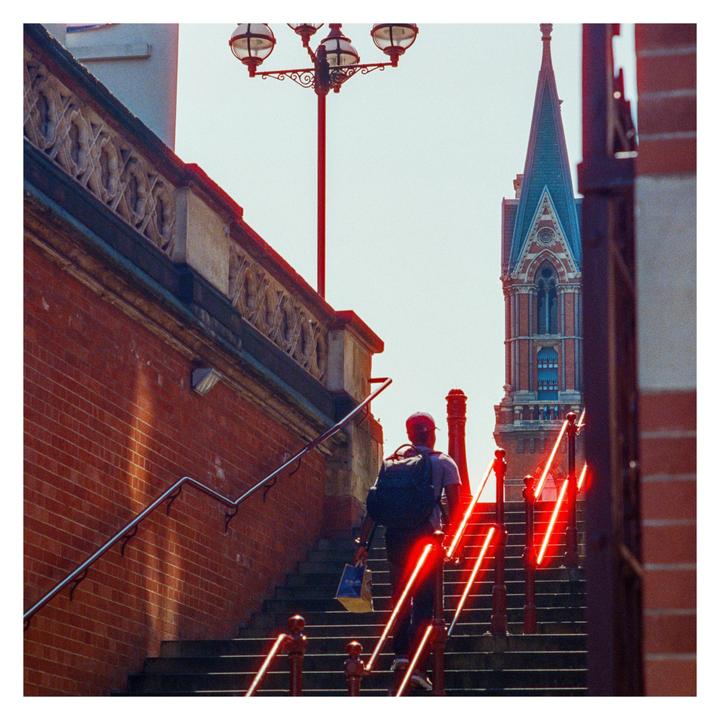 A man with a backpack walks up a set of stairs with red light trails, towards a tall building with a spire.