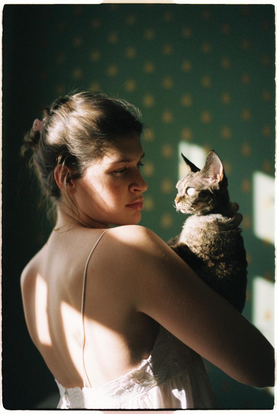 A woman holds a cat in her arms, looking at it with soft light falling on her back and the cat.