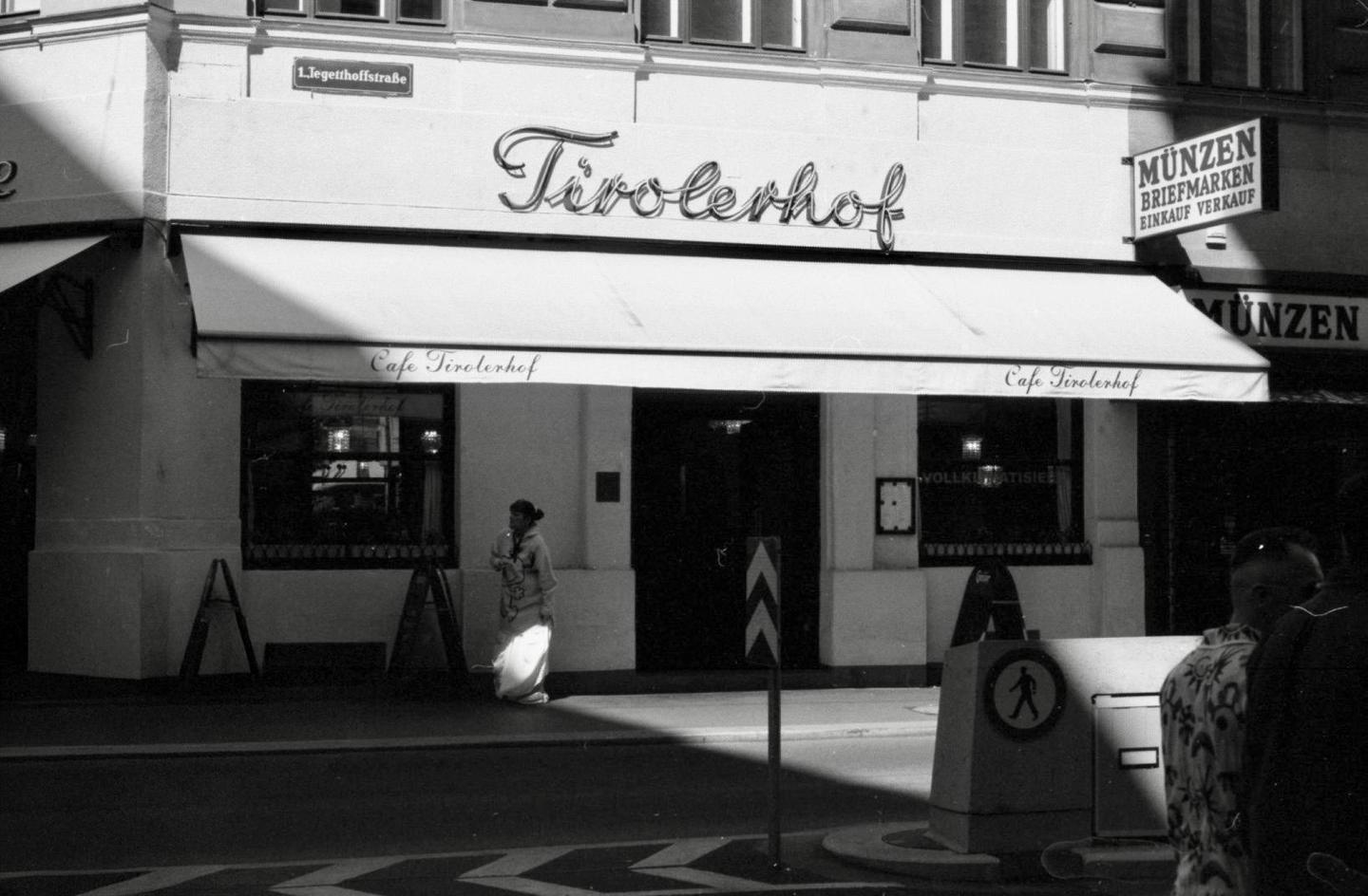 A woman stands on a sunlit sidewalk in front of a cafe with an awning and a neon sign.