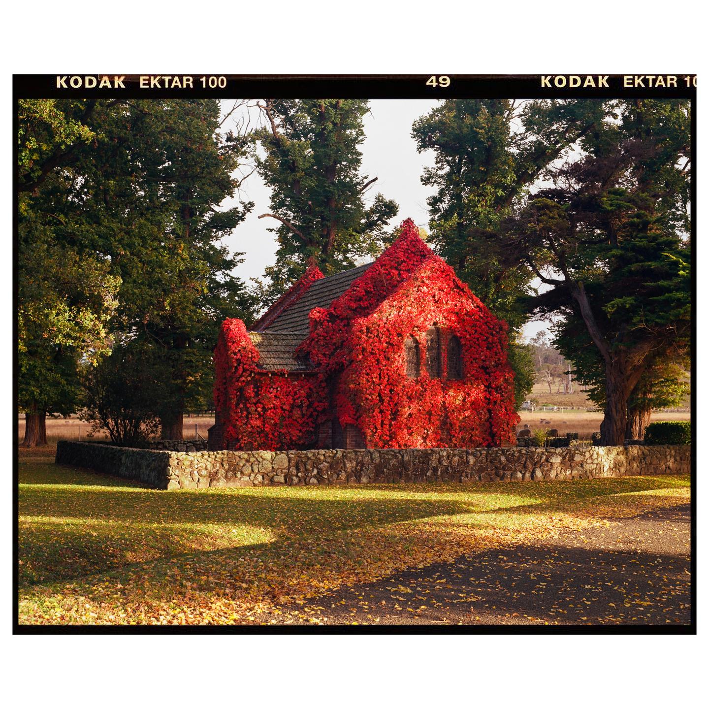 A small church covered in red ivy stands behind a stone wall with trees and fallen leaves in the foreground.