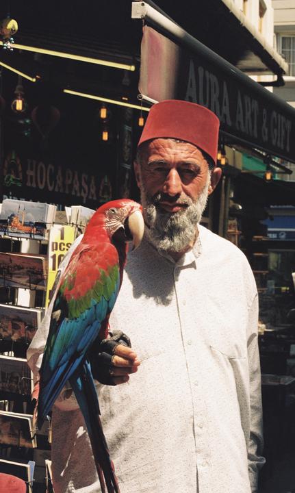 A man wearing a red fez and a white shirt holds a colorful parrot on his gloved hand.