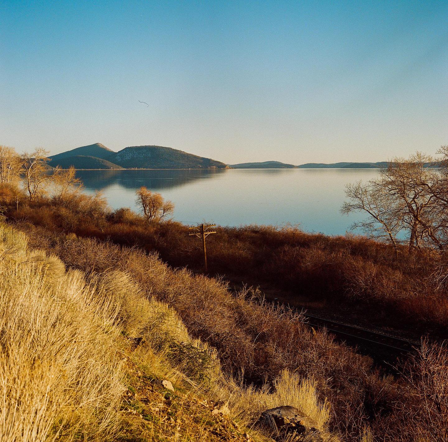 A body of water reflects mountains under a clear sky, with dry brush and a telephone pole in the foreground.