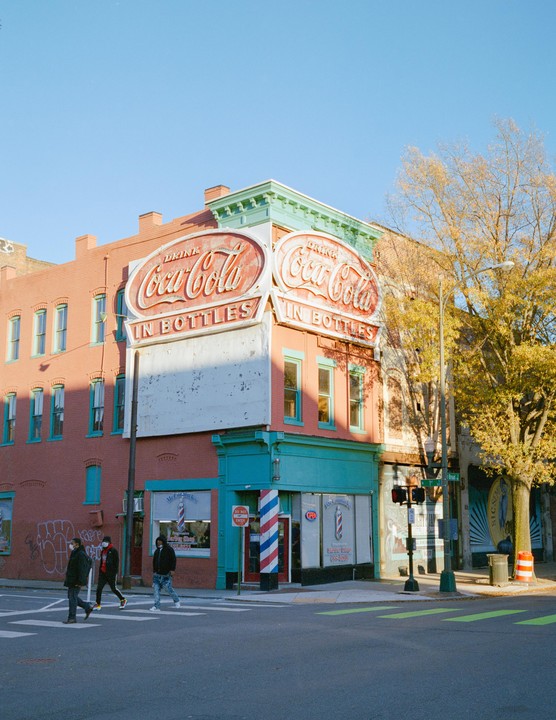 Three men cross a street in front of a brick building with two large Coca-Cola signs.