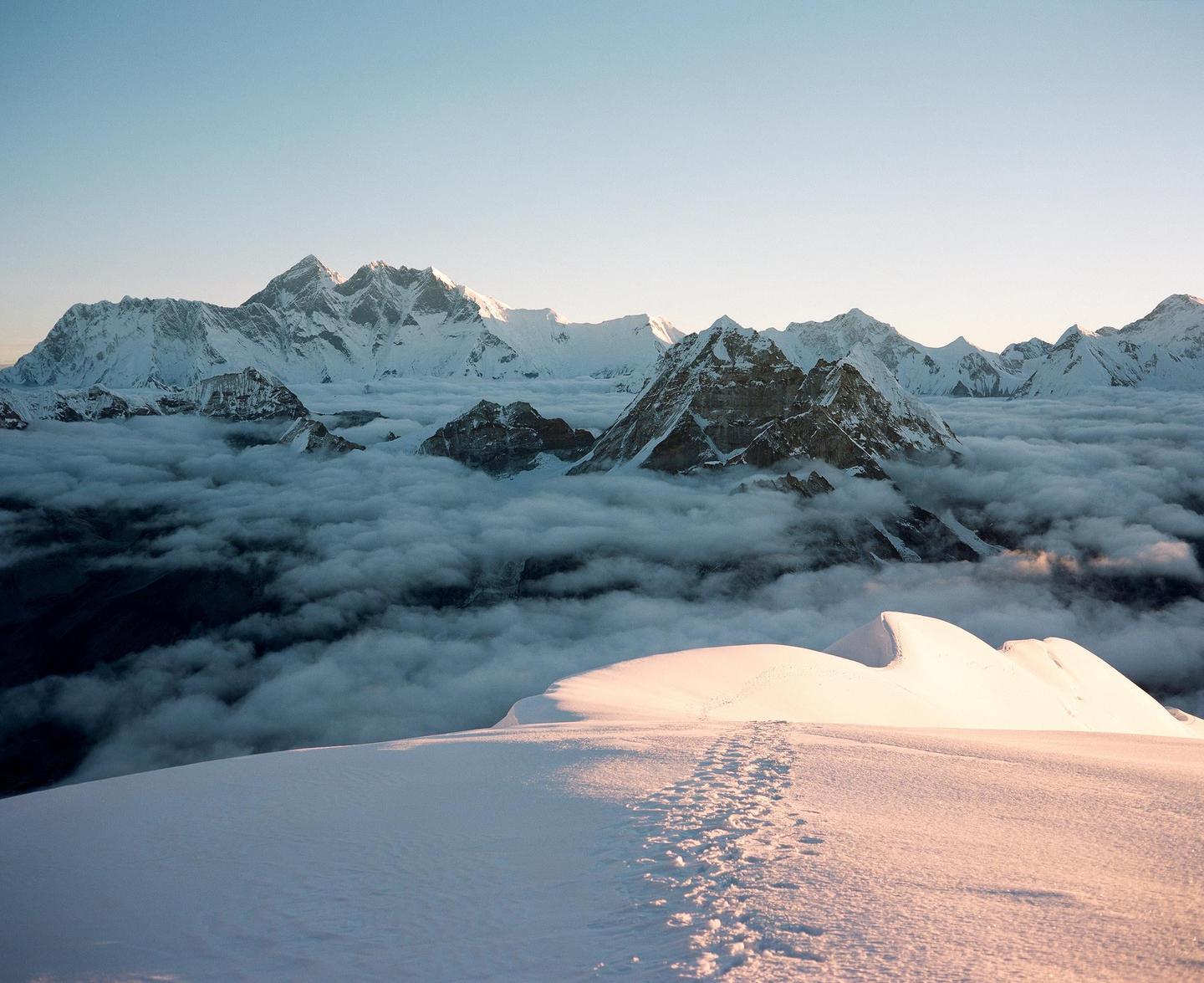 Snow-covered mountains rise above a sea of clouds with footprints visible in the foreground.