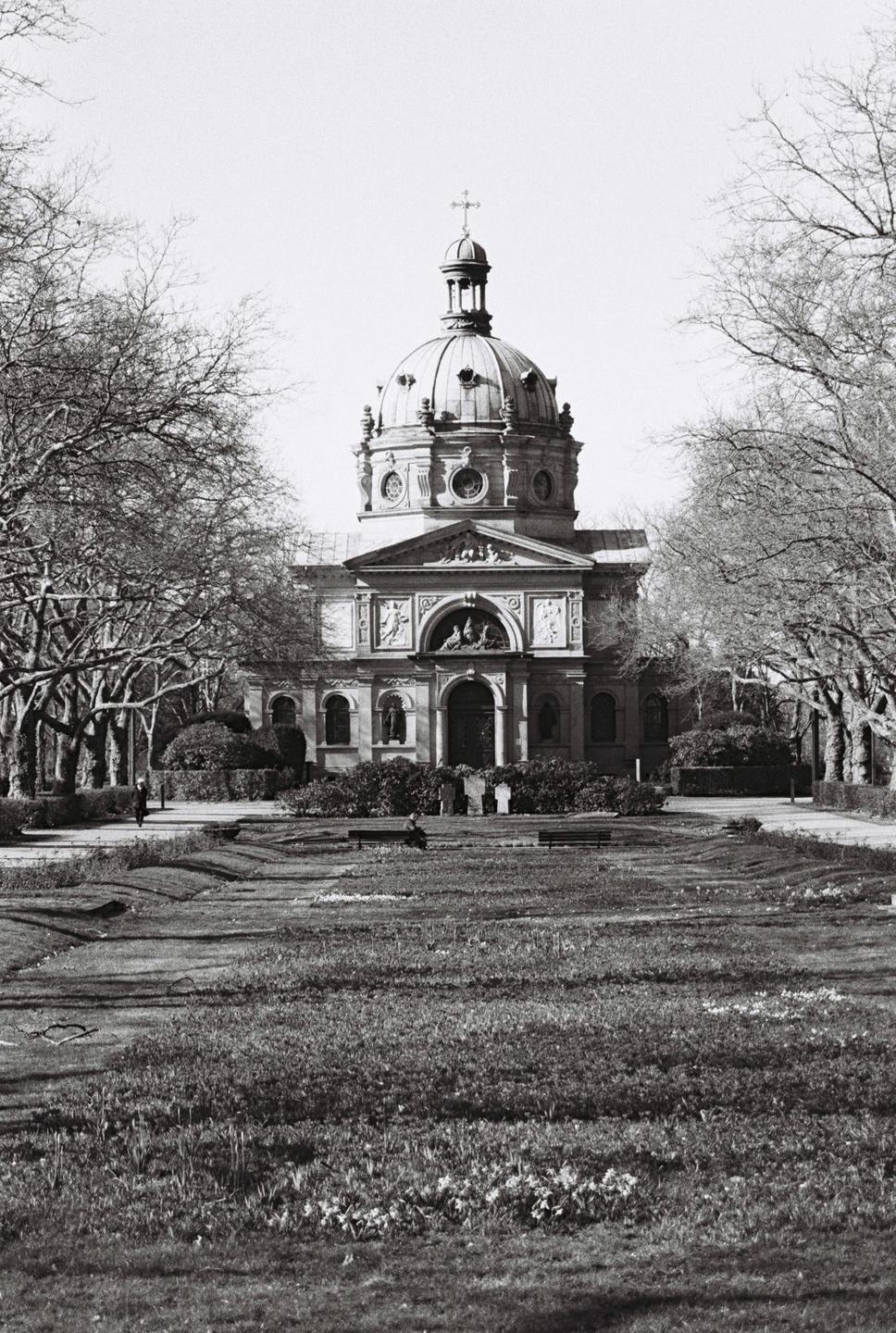 A domed building with a cross on top stands at the end of a grassy area lined with trees.