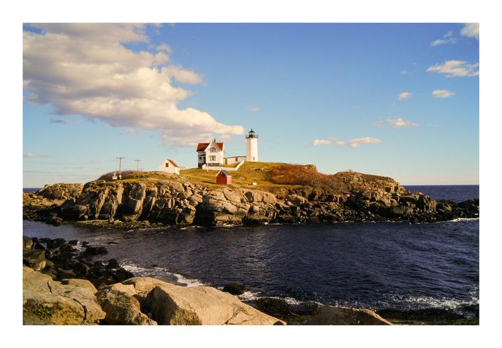 A white lighthouse and buildings stand on a rocky island surrounded by dark blue water under a partly cloudy sky.