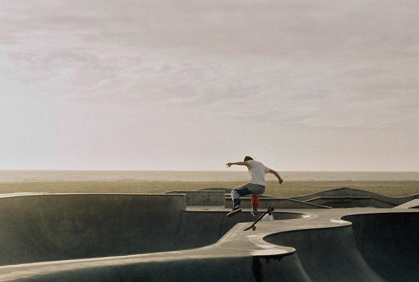 A man on a skateboard is captured mid-air over a skatepark with the ocean in the background.