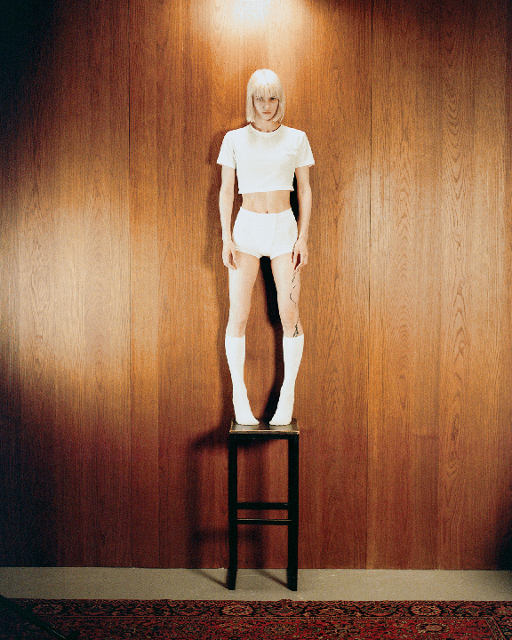 A woman stands on a stool in front of a wood-paneled wall, wearing a white crop top, white shorts, and white socks.