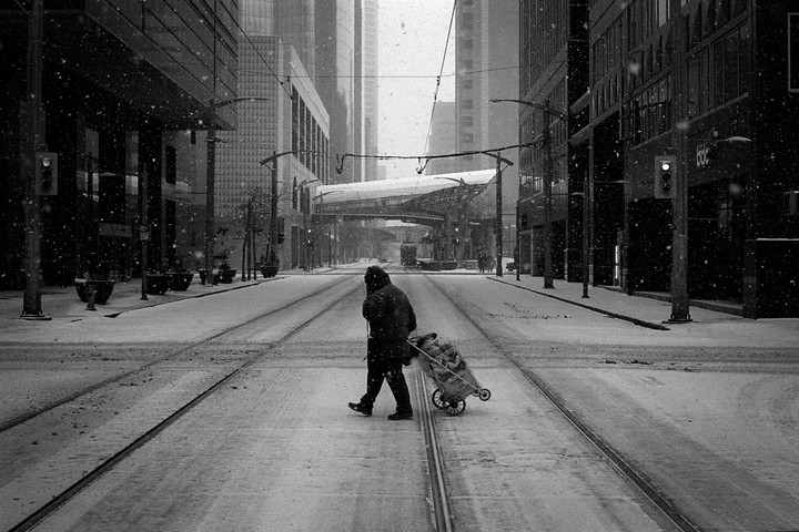 A person pulls a cart across a snowy street with tram tracks, surrounded by tall buildings.