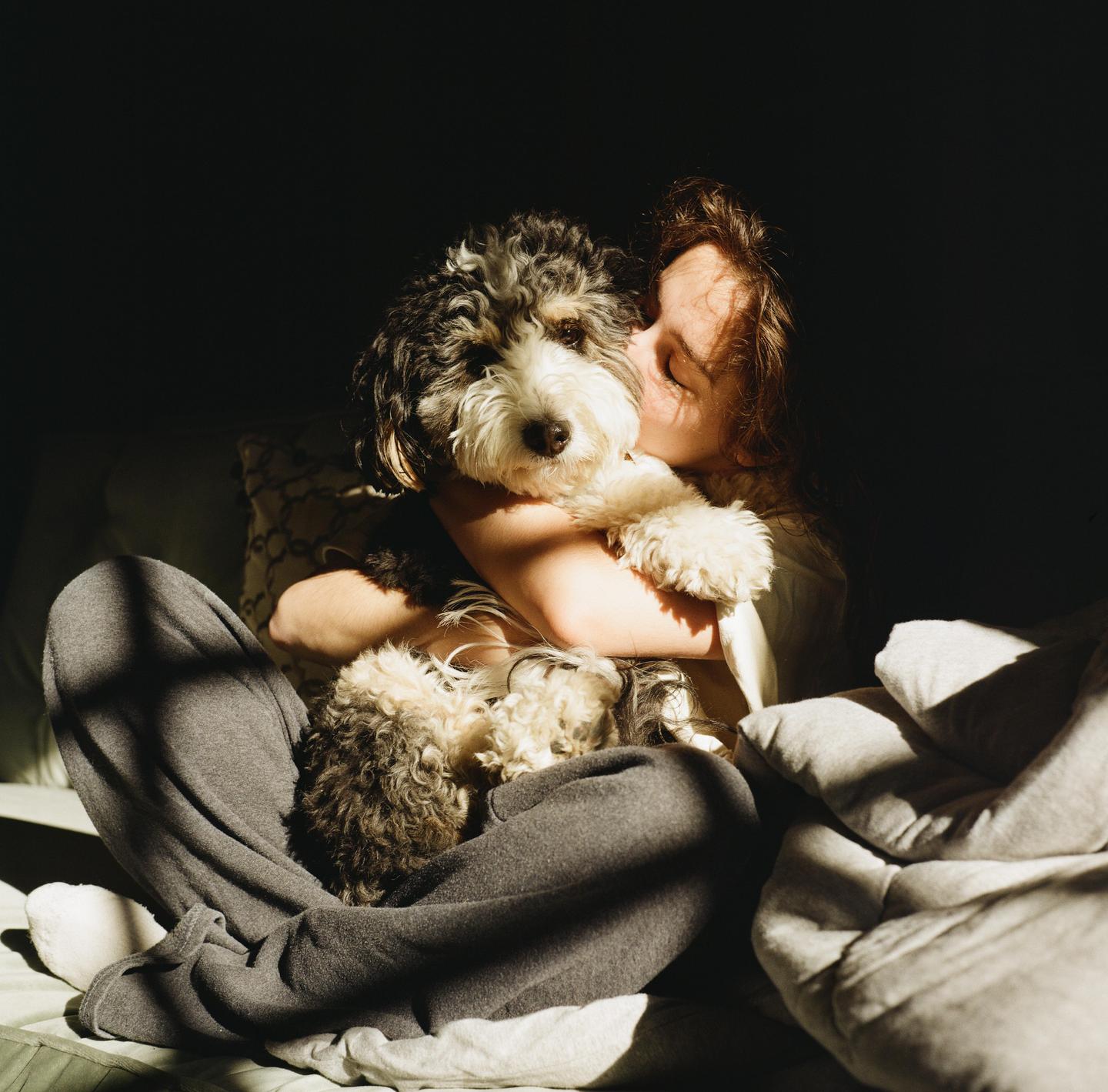 A woman sits on a bed, hugging a fluffy dog in a sunlit room.