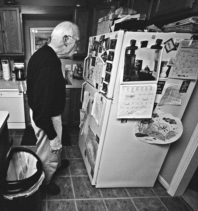 A man stands in a kitchen looking at a refrigerator covered with magnets and papers.