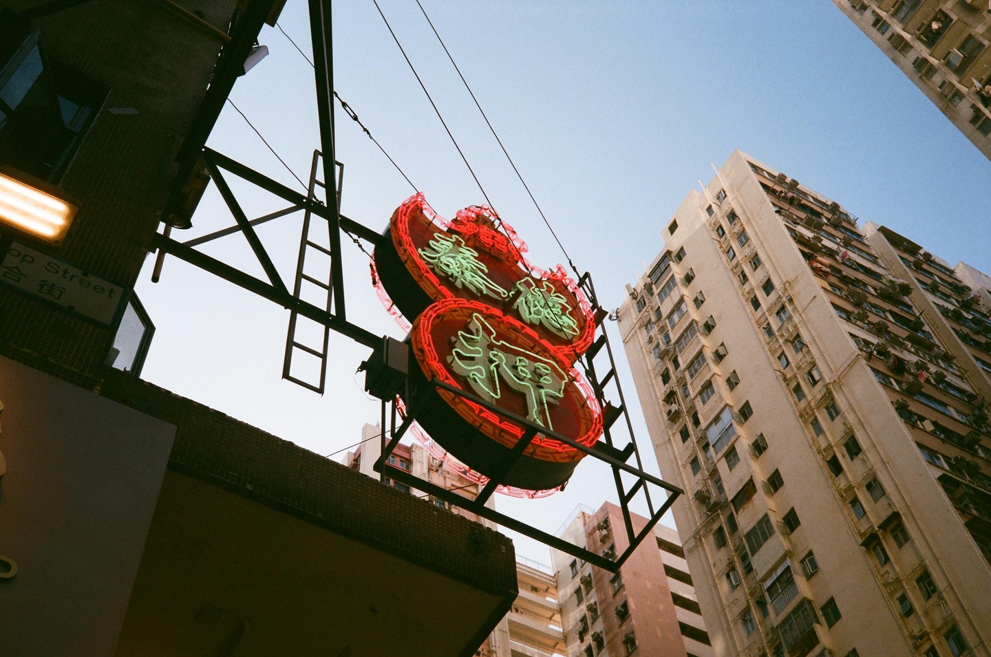 A neon sign with red and green lights hangs above a street with tall buildings under a clear sky.