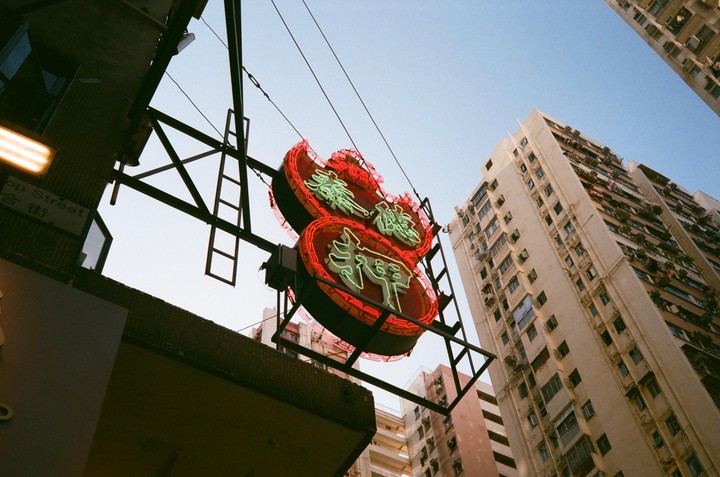 A neon sign with red and green lights hangs above a street with tall buildings under a clear sky.