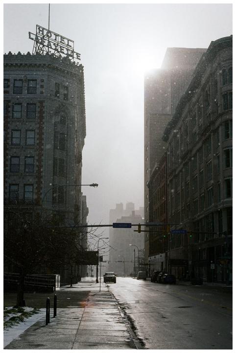 A street in a city is covered in snow with tall buildings on either side and a bright sky.