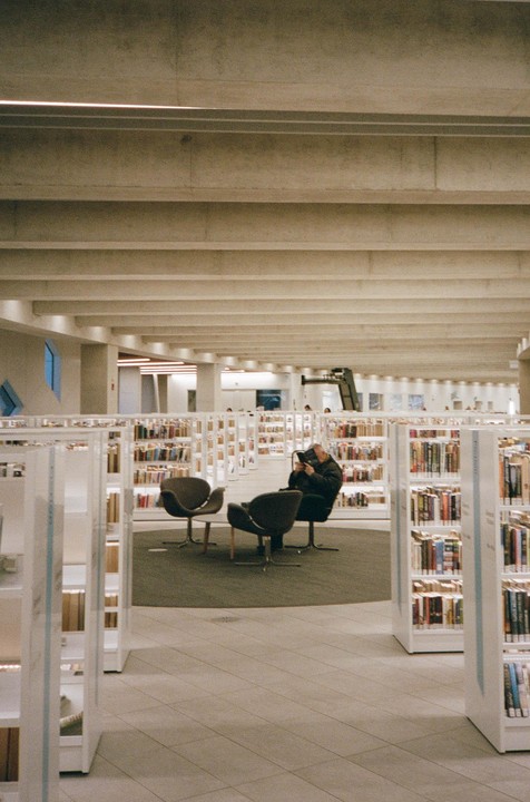 A man sits in a chair reading a book in a library surrounded by bookshelves.