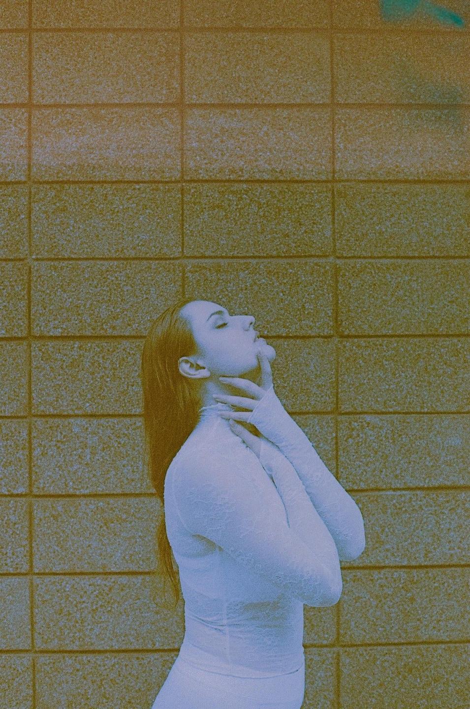 A woman in a white lace garment poses with her head tilted back and her hands on her neck against a textured wall.
