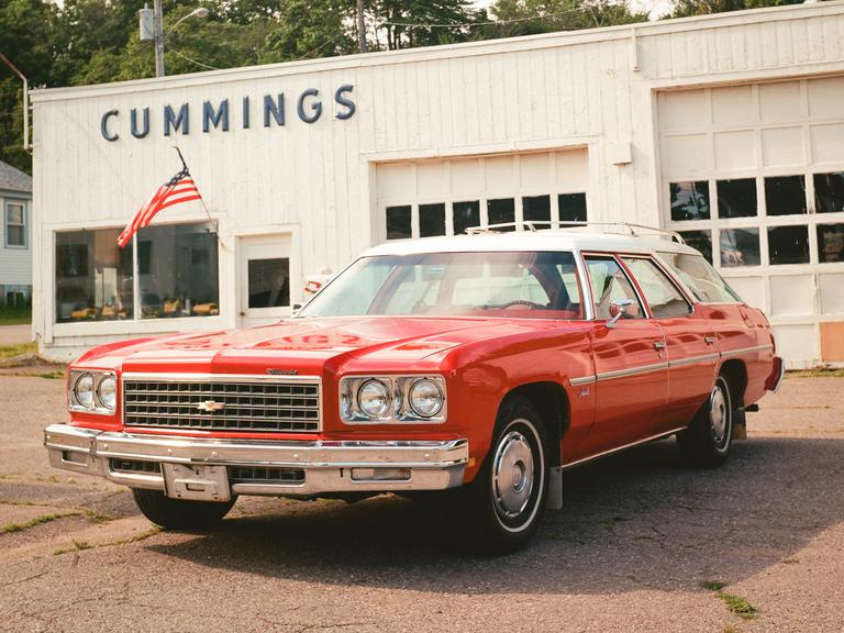 A red station wagon is parked in front of a white building with the name "CUMMINGS" on it.