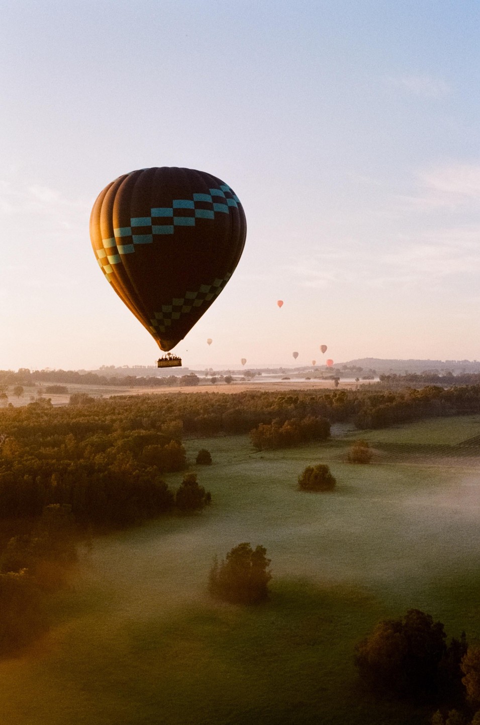 A hot air balloon with a checkered pattern floats above a misty landscape with trees and fields during sunrise.