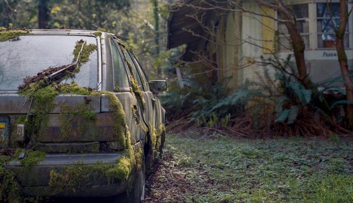 An abandoned car covered in moss is parked in a forest next to a house.