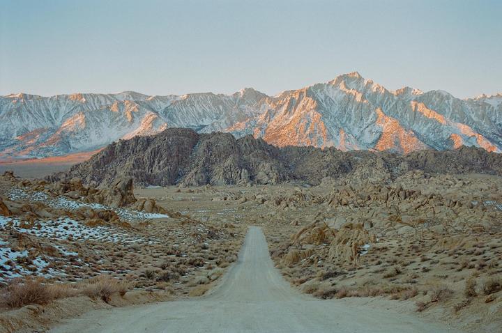 A dirt road leads toward snow-capped mountains with rocky hills in the foreground.