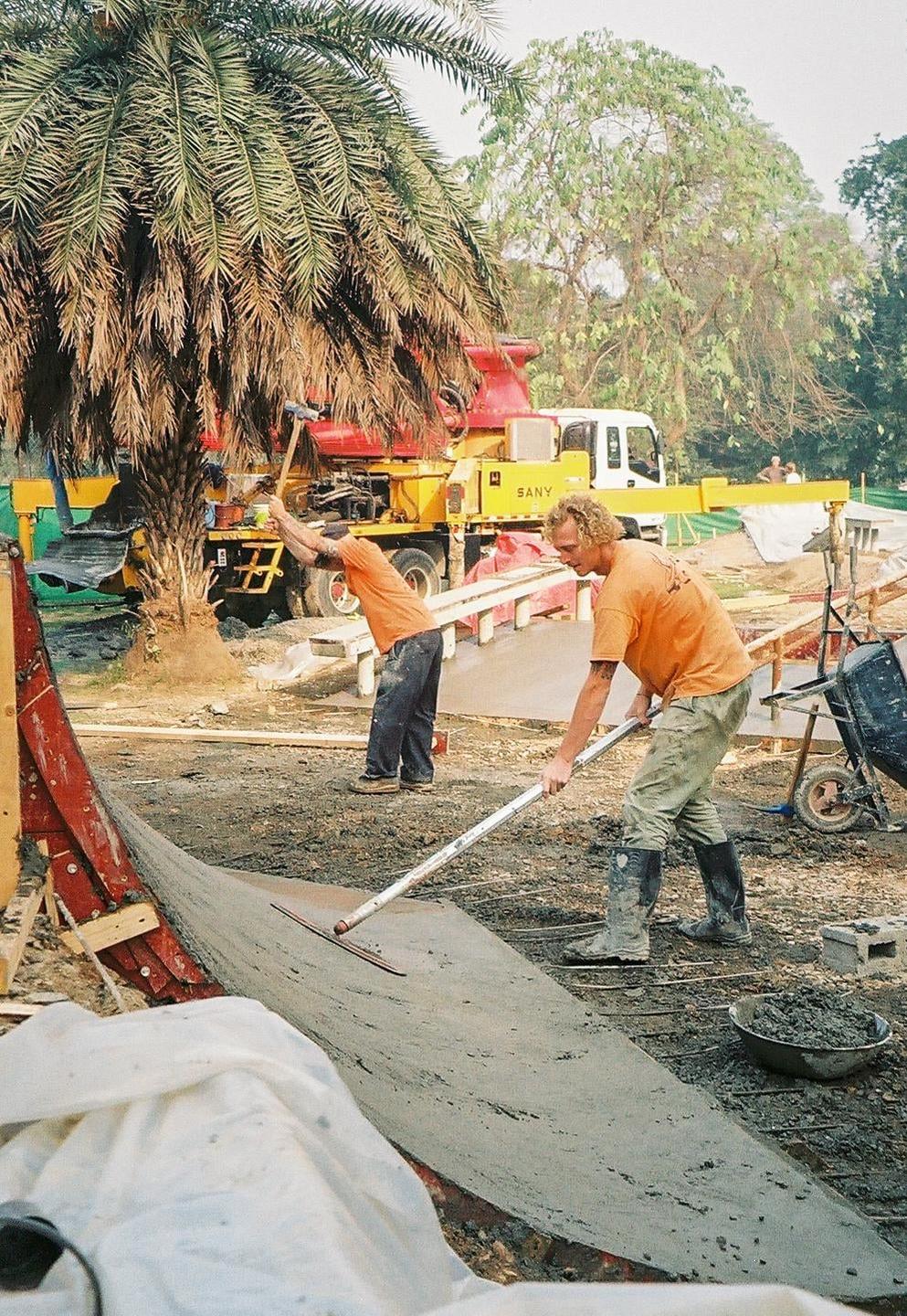 Two men are working on a concrete skatepark ramp next to a palm tree and construction vehicles.