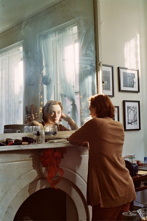 A woman in a brown cardigan leans on a fireplace mantel, looking at her reflection in a large mirror.