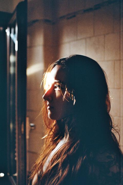 A woman with long hair and a nose piercing is illuminated by a shaft of light from the left.