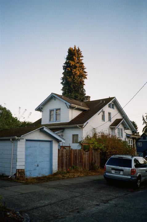 A white house with a blue garage and a silver minivan parked on the street in front of a wooden fence.