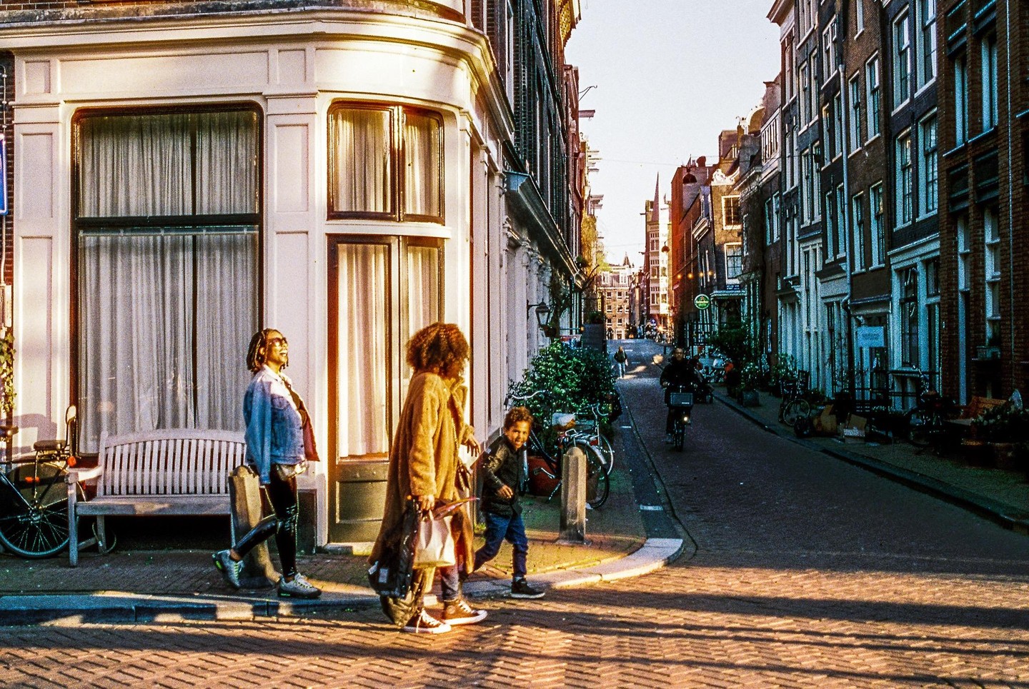 Three people walk on a sunlit street corner with tall buildings and bicycles lining the street.