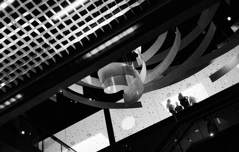 A group of people stand on a balcony overlooking a modern interior with a patterned ceiling and hanging decorations.