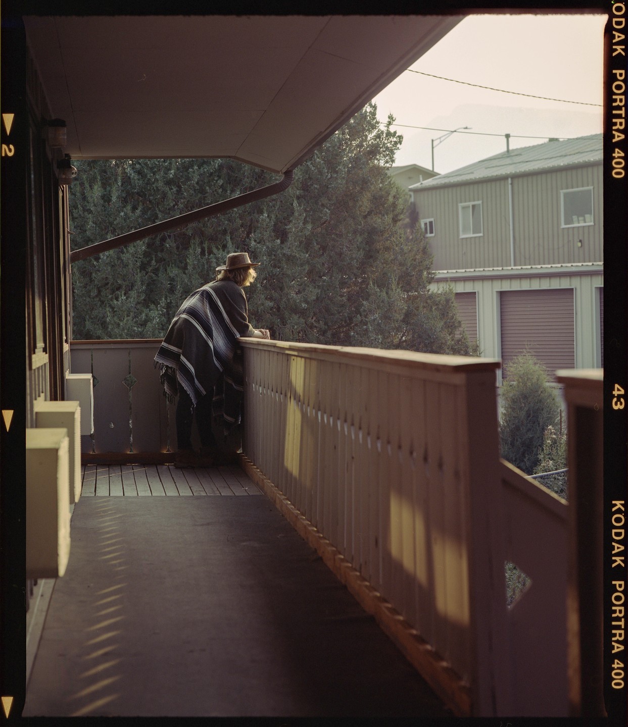 A person wearing a poncho and hat leans on a balcony railing, looking out at a hazy landscape.