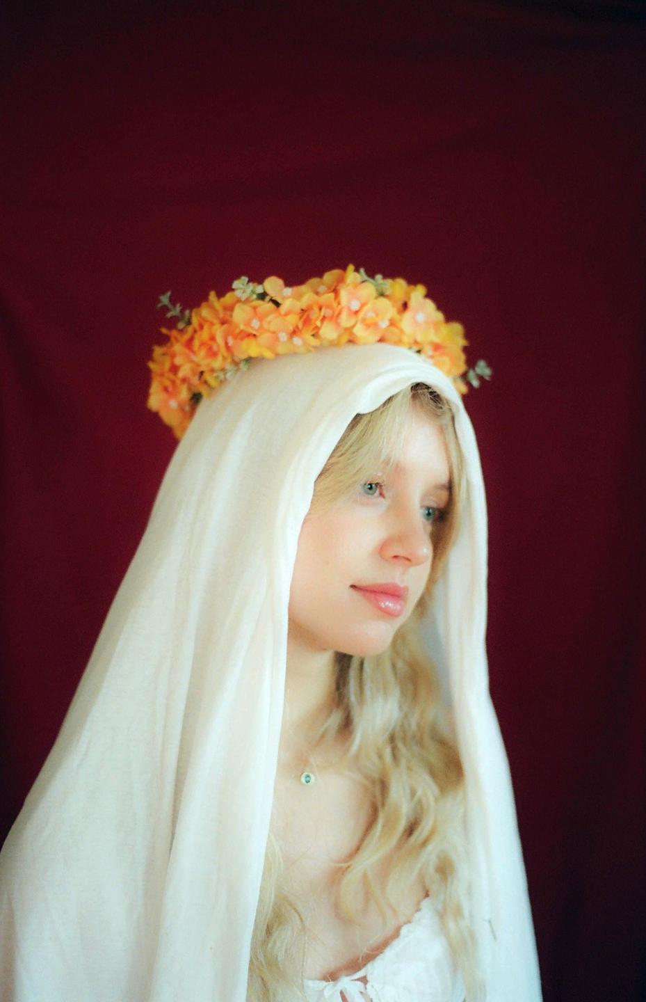 A woman with long blonde hair and a white veil wears a flower crown and looks to the right.